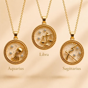 May include: Gold-toned necklaces featuring zodiac symbols: Aquarius, Libra, and Sagittarius. Each pendant has a round design with a textured border, a constellation of small crystals, and a central zodiac symbol. The word "Libra", "Aquarius", and "Sagittarius" are printed on the image.