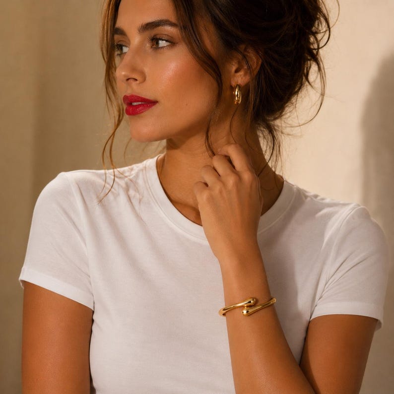 May include: A gold-toned metal cuff bracelet with a smooth, polished finish and rounded, tapered ends, worn on the wrist. The bracelet features a minimalist open design, complemented by a pair of small, matching gold-toned hoop earrings.