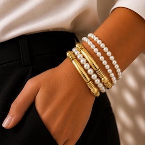 Op de afbeelding: Een set van vier armbanden die samen worden gedragen. Twee armbanden bestaan uit kleine witte bolvormige kralen. De andere twee armbanden bevatten een combinatie van kleine witte kralen en gladde, cilindrische goudkleurige metalen staven.
