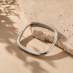 May include: A polished silver-tone metal bangle bracelet with a distinct square shape and rounded corners. The bracelet features a smooth, reflective surface and a visible hinge mechanism on one side, allowing it to open and close securely.