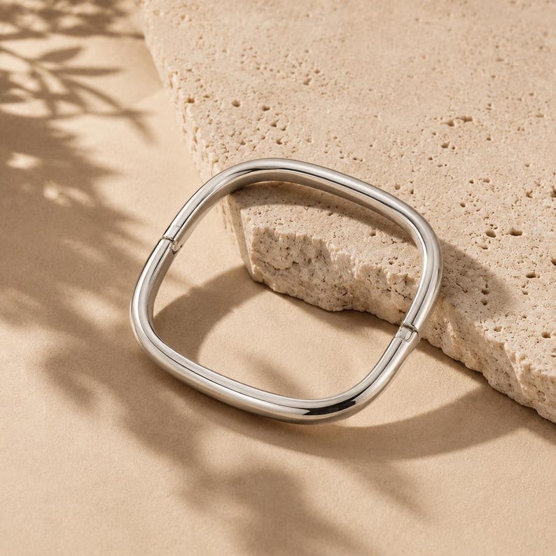 May include: A polished silver-tone metal bangle bracelet with a distinct square shape and rounded corners. The bracelet features a smooth, reflective surface and a visible hinge mechanism on one side, allowing it to open and close securely.