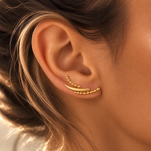 Pode incluir: Um brinco trepador dourado com uma barra central curva e lisa, ladeada por duas filas de pequenas contas esféricas. A peça de joalharia segue a curva natural do lóbulo e da cartilagem da orelha, criando um aspeto em camadas e texturado.