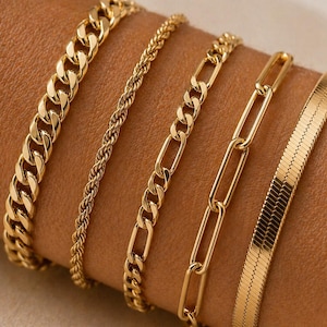 Peut inclure: Cinq bracelets dorés de différents modèles sont présentés. Les bracelets comprennent une chaîne gourmette, une chaîne torsadée, une chaîne Figaro, une chaîne trombone et une chaîne chevrons. Les bracelets sont présentés sur le bras d'une personne.