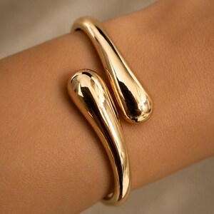 May include: A polished gold-tone cuff bracelet featuring two smooth, teardrop-shaped ends that overlap slightly. The metal surface is highly reflective, showcasing a curved, fluid design that wraps around the wrist with a sleek, continuous form.
