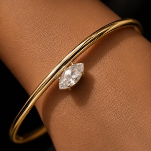 May include: A polished gold bangle bracelet featuring a single marquise-cut clear gemstone set in a prong setting. The smooth, rounded metal band encircles the wrist, showcasing a minimalist design with a central, faceted stone accent.