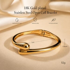 May include: An 18K gold plated stainless steel open cuff bracelet with a polished finish. The text reads 18K Gold plated Stainless Steel Open Cuff Bracelet, Water Resistant, Tarnish Free, Hypoallergenic. It measures 7cm by 0.8cm and weighs 52g.