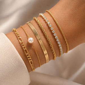 May include: A collection of gold bracelets on a wrist. The bracelets include a chain link, a delicate chain with a pearl, a herringbone design, a twisted rope style, and a tennis bracelet with clear stones.