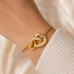 May include: A gold-colored bracelet featuring two interlocking heart pendants. One heart is encrusted with small, clear gemstones. The bracelet has a delicate chain and is worn on a wrist, suggesting a jewelry item.