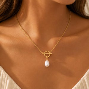 Puede incluir: Un collar dorado con un cierre circular y un colgante de perla blanca. El collar se lleva alrededor del cuello, mostrando la fina cadena y el diseño elegante. La perla es una adición clásica y atemporal.