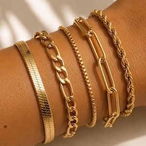 Puede incluir: Colección de cinco pulseras doradas con diferentes diseños. Las pulseras incluyen una espiga, una cadena de eslabones, una cadena de caja, una cadena de clip y una cadena de cuerda. Las pulseras se muestran en una muñeca.