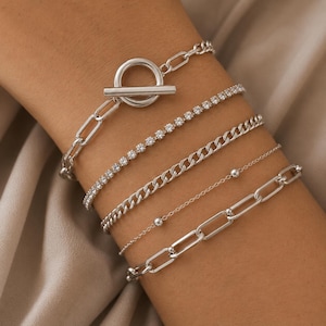 Può includere: Un set di cinque bracciali color argento in vari stili, tra cui una catena con chiusura a T, una catena tennis con cristalli incastonati, una catena a maglia gourmette spessa, una catena sottile con perline e una catena a maglie lunghe.