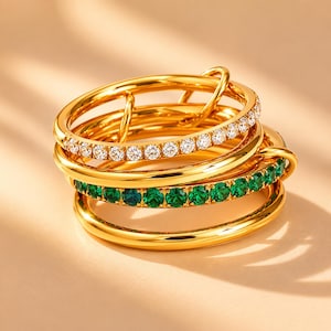 May include: A stack of four gold-toned rings. The top ring features a row of clear, round-cut stones. The third ring from the top features a row of round-cut green stones. The other two rings are plain, polished gold-toned bands with smooth surfaces.