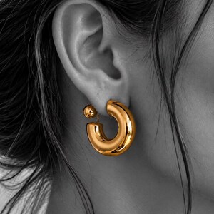 May include: A gold-toned hoop earring with a thick, rounded, polished surface. The earring features a small, spherical ball stud detail that sits behind the earlobe, connected to the main hoop by a thin post, creating a layered, dimensional look.