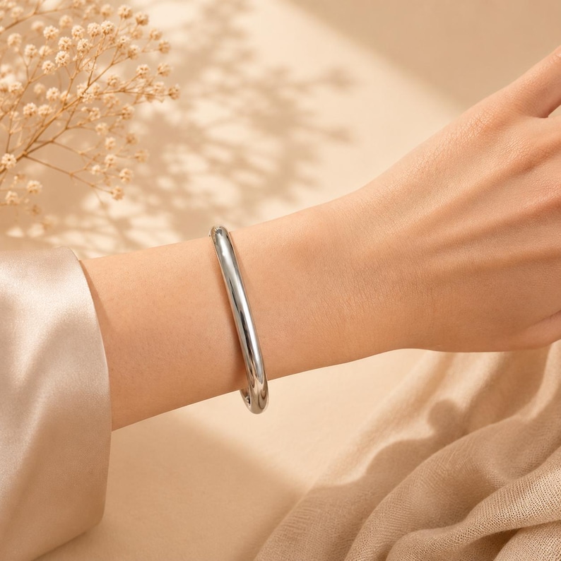 May include: A smooth, polished silver-tone bangle bracelet with a rounded, tubular profile. The bracelet features a continuous, seamless design that encircles the wrist, showcasing a reflective metallic surface that catches the light.
