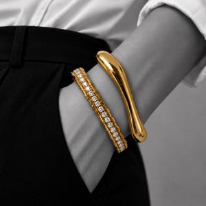 Peut inclure: Deux bracelets dorés portés ensemble. L'un est un jonc rigide, lisse et incurvé avec une finition polie. Le second est un bracelet à maillons souples doté d'une rangée centrale de petites pierres rondes transparentes serties dans une monture dorée.