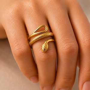 May include: A gold-toned ring shaped like a coiled snake. The band features a textured, scale-like pattern along the body of the snake, with a smooth, looped tail end and a detailed, rounded head that rests against the finger, creating a wrap design.