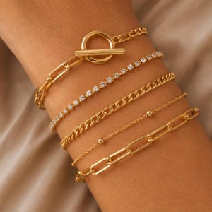 Könnte beinhalten: Eine Auswahl an goldfarbenen Armbändern. Die Armbänder umfassen eine Kettenglied-Variante mit Knebelverschluss, ein Tennisarmband mit klaren Steinen, eine Panzerkette, eine zarte Perlenkette und eine Büroklammerkette.