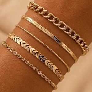 May include: A collection of gold-colored bracelets. The bracelets include a chunky chain link, a flat snake chain, a thin cable chain, a chevron design, and a rope chain. The bracelets are stacked on a wrist.