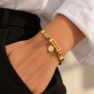 May include: A gold-toned metal cuff bracelet featuring a central bar design with two spherical accents on either side and a small, circular dangling charm attached to the middle section, creating a polished and reflective metallic appearance.