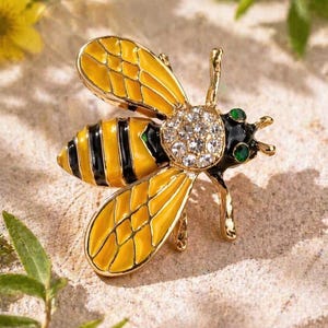 May include: A gold-coloured bee brooch featuring yellow enamel wings and black stripes on the abdomen. The bee's head is decorated with clear crystals and green accents. The brooch is set against a neutral background.