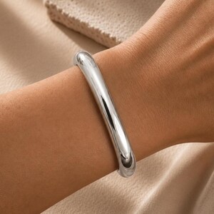 May include: A polished silver-tone bangle bracelet with a smooth, rounded, and continuous tubular design. The metal surface is highly reflective, showing bright highlights and deep shadows that emphasize its curved, solid, and seamless form.