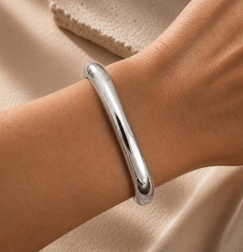 May include: A polished silver-tone bangle bracelet with a smooth, rounded, and continuous tubular design. The metal surface is highly reflective, showing bright highlights and deep shadows that emphasize its curved, solid, and seamless form.