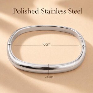 May include: A polished stainless steel bangle bracelet with a smooth, rounded, oval shape. The bracelet features a hinged opening mechanism. Text on the image indicates a width of 6cm and a thickness of 0.85cm for the metal band.