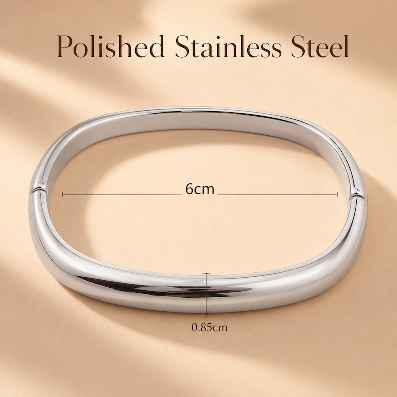 May include: A polished stainless steel bangle bracelet with a smooth, rounded, oval shape. The bracelet features a hinged opening mechanism. Text on the image indicates a width of 6cm and a thickness of 0.85cm for the metal band.