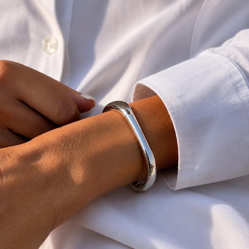 May include: A polished silver-tone bangle bracelet with a smooth, rounded, and slightly irregular organic shape. The bracelet features a continuous, solid metal band that encircles the wrist, reflecting light with a high-shine, reflective finish.