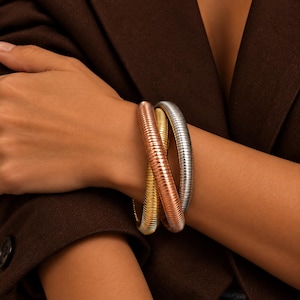 May include: A set of three interlocking bangle bracelets featuring a flexible, ribbed, snake-like texture. The bracelets are finished in metallic gold, copper, and silver tones, creating a layered appearance around the wrist.