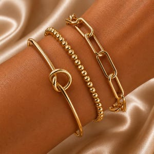 May include: Three gold-colored bracelets are displayed on a wrist. The bracelets include a knot-shaped bangle, a beaded bracelet, and a chain link bracelet. The bracelets are made of a shiny, metallic material.