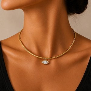 May include: A gold-toned metal choker necklace featuring a smooth, rigid circular band design. A single marquise-cut clear gemstone pendant is suspended from the centre of the necklace, held by a small gold-toned metal loop and setting.