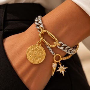 May include: A mixed-metal chain bracelet featuring a gold-tone carabiner clasp, a round gold-tone medallion with an embossed cross design, a small gold-tone cone charm with clear stones, and a gold-tone star charm with clear stones.