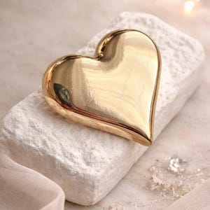 May include: A polished, gold-colored heart-shaped object with a smooth, reflective surface. The heart is positioned on a textured, off-white rectangular block, accompanied by small, clear, faceted decorative stones and a light-colored fabric.