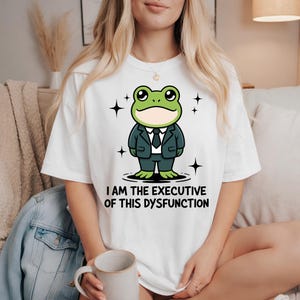 Könnte beinhalten: Weißes T-Shirt mit einer Cartoon-Froschfigur im Anzug und dem Text "I AM THE EXECUTIVE OF THIS DYSFUNCTION". Der Frosch ist grün und trägt einen schwarzen Anzug und eine Krawatte. Schwarze Sterne umgeben den Frosch.
