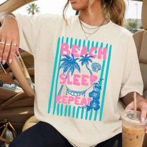 May include: Cream-colored t-shirt with a tropical graphic. The design features the words "BEACH SLEEP REPEAT" in pink, with blue palm trees, a seashell, and a hammock. The shirt has a relaxed fit.