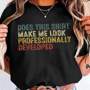 Peut inclure: Un t-shirt noir à manches courtes avec un texte multicolore qui indique : DOES THIS SHIRT MAKE ME LOOK PROFESSIONALLY DEVELOPED. Le lettrage est disposé sur quatre lignes avec une police de style vintage usé dans des tons bleu sarcelle, jaune et rouge.