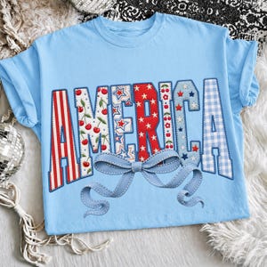May include: Light blue t-shirt with the word "AMERICA" in a patchwork design. The letters feature red and white stripes, cherries, stars, and a blue gingham pattern. A light blue bow is stitched below the word.