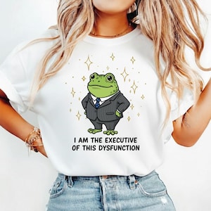 Peut inclure: T-shirt blanc avec une grenouille de dessin animé en costume et cravate, avec le texte "I AM THE EXECUTIVE OF THIS DYSFUNCTION". La grenouille est verte, le costume est gris, et des éclats d'étoiles dorées entourent la grenouille.