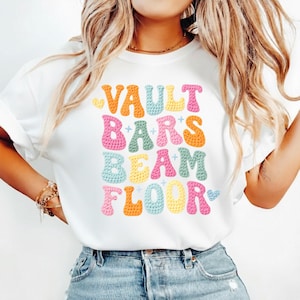 Puede incluir: Camiseta blanca con las palabras "VAULT BARS BEAM FLOOR" en letras de burbujas coloridas. El texto está dispuesto verticalmente, con cada palabra en un color diferente, incluyendo naranja, rosa, azul, amarillo y un toque de azul claro. La camiseta es de estilo casual.