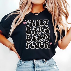 Puede incluir: Camiseta negra con las palabras "VAULT BARS BEAM FLOOR" en una fuente retro audaz. El texto está delineado en blanco y tiene una apariencia texturizada, similar al ganchillo. Acentos de corazón rosa se colocan en la parte superior e inferior del texto.