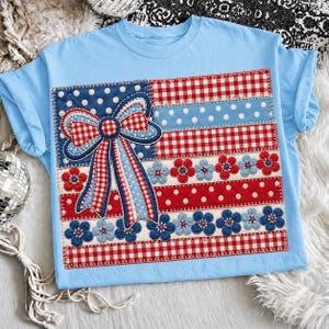 Può includere: T-shirt azzurra con un design patriottico. Il design include un grande fiocco, pois, quadretti e accenti floreali in rosso, bianco e blu. La maglietta è realizzata in materiale morbido.