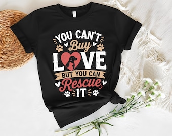 Peut inclure: T-shirt noir avec l'inscription "YOU CAN'T BUY LOVE BUT YOU CAN RESCUE IT." Le motif comprend un cœur avec une silhouette de chien, des empreintes de pattes et des éléments décoratifs blancs, rouges et bruns.