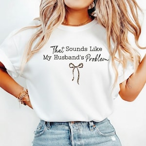May include: White t-shirt with the text "That Sounds Like My Husband's Problem" in black script. A leopard print bow graphic is below the text. The shirt is paired with blue jeans.