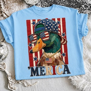 Può includere: T-shirt azzurra con un'immagine di un'anatra patriottica con occhiali da sole, berretto da baseball e una catena d'oro. L'anatra è posta su uno sfondo a strisce rosse e bianche con stelle, e la parola "MERICA" è visualizzata sotto.