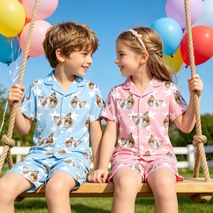 Könnte beinhalten: Zwei Kinder in passenden Pyjama-Sets. Das eine Set ist hellblau mit Katzenköpfen und weißen Sternen, das andere ist rosa mit dem gleichen Katzen- und Sternmuster. Sie sitzen auf einer Holzschaukel, lächeln, mit bunten Luftballons im Hintergrund.