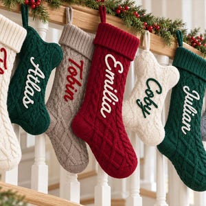 Peut inclure: Une rangée de sept chaussettes de Noël en tricot torsadé suspendues à une rampe en bois. Les chaussettes sont crème, vert foncé, beige, rouge et gris, chacune portant un nom brodé dans une couleur contrastante : Florence, Atlas, Tobin, Emilia, Coby, Julian et Niko.