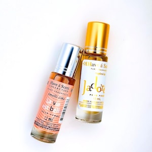 Puede incluir: Dos frascos de perfume sobre una superficie blanca. Un frasco es melocot&oacute;n con tapa plateada y texto "La vie est belle" y "12 ml". El otro frasco es transparente con tapa dorada y texto "J'adore" y "12 ml".