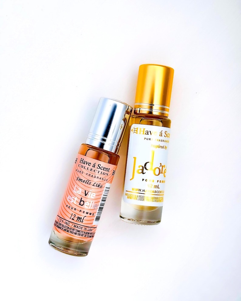 Puede incluir: Dos frascos de perfume sobre una superficie blanca. Un frasco es melocot&oacute;n con tapa plateada y texto "La vie est belle" y "12 ml". El otro frasco es transparente con tapa dorada y texto "J'adore" y "12 ml".