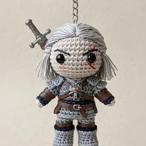 May include: Crocheted keychain featuring a doll with gray hair and a scar, dressed in gray armor with brown accents. A silver sword is attached. The doll has a silver keychain ring.